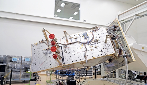 The JPSS-4 spacecraft appears as a silver rectangle in the center of the image with wires and hardware attached to the silver panel. The spacecraft is attached to a white, rotating fixture and sits in a white room with an observation window in the top, center of the image. The room also contains metal crates, a desk, a chair, trash can, and metal stepping stool.