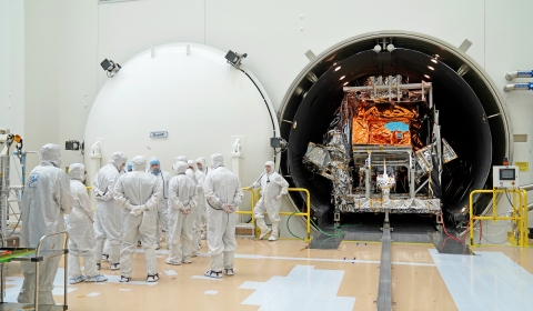 In a large white clean room with a tan and white floor, the JPSS-3 spacecraft is seen inside of a dark, tubular chamber with rows of lights on the ceiling. A large, circular door is open to the left. The spacecraft is covered in silver and orange colored mylar. On the left, in front of the chamber door, a group of 12 individuals are standing and talking while wearing white bunny suits. Yellow railing sits on both sides of the chamber opening.