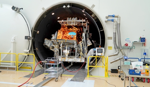 A large, tubular champer sits open in a large, white clean room with its white door seen on the left against a white walll. The silver and orange mylar-covered JPSS-3 satellite sits inside the chamber while a person in a white bunny suit works on the right side of the spacecraft. Outside of the chamber there is yellow railing on both sides of the tube.  There are silver pipes attached to the wall next to the chamber opening. Outside of the chamber, there is a tan and white floor, silver carts and tables wit