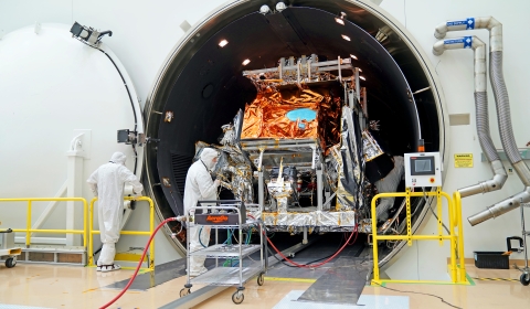 A large, tubular chamber sits open in a large, white clean room with its white door seen on the left against a white wall. The silver and orange mylar-covered JPSS-3 satellite sits inside the chamber while two individuals work in front of the chamber. Yellow railing sits on both sides of the chamber opening. Silver pipes are attached to the wall on the right side of the chamber. A silver cart with red wires attached sits directly in front of the spacecraft.