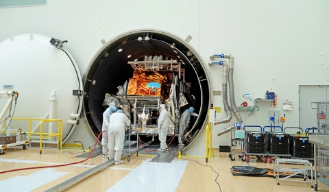 A large, tubular chamber sits open in a large, white clean room with its white door seen on the left against a white wall. The silver and orange mylar-covered JPSS-3 satellite sits inside the chamber while five individuals in white bunny suits work around the spacecraft. Three people are directly in front of the spacecraft around a silver cart with red wires attached. One stands in front of the spacecraft to the right side and one other is working inside the chamber on the right side.