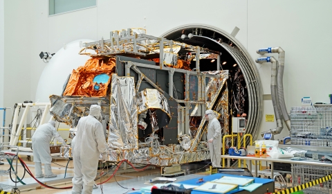 In the center of the photo, the spacecraft covered in silver and orange mylar sits just outside of a large, tubular chamber. Three individuals in white bunny suits work on the spacecraft in a large, white clean room. Silver pipes are attached to the wall to the right side of the open chamber and on the left side, the open white door to the chamber is visible. Yellow railing sits on both side of the chamber. In the foreground of the photo, yellow and black tape hangs to close off the area and a desk.