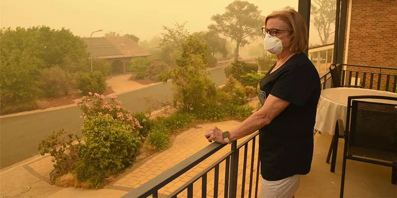 The image depicts a woman standing on a balcony wearing a protective face mask. She is looking off into the distance, where a thick, orange haze from a nearby wildfire obscures the view of the surrounding area. The haze gives the scene an eerie, sepia-toned appearance. The woman is dressed casually in a black t-shirt with a circular design and glasses, and she is wearing a watch on her left wrist. She appears contemplative or concerned as she gazes at the smoky landscape. There's a quiet, almost desolate fe
