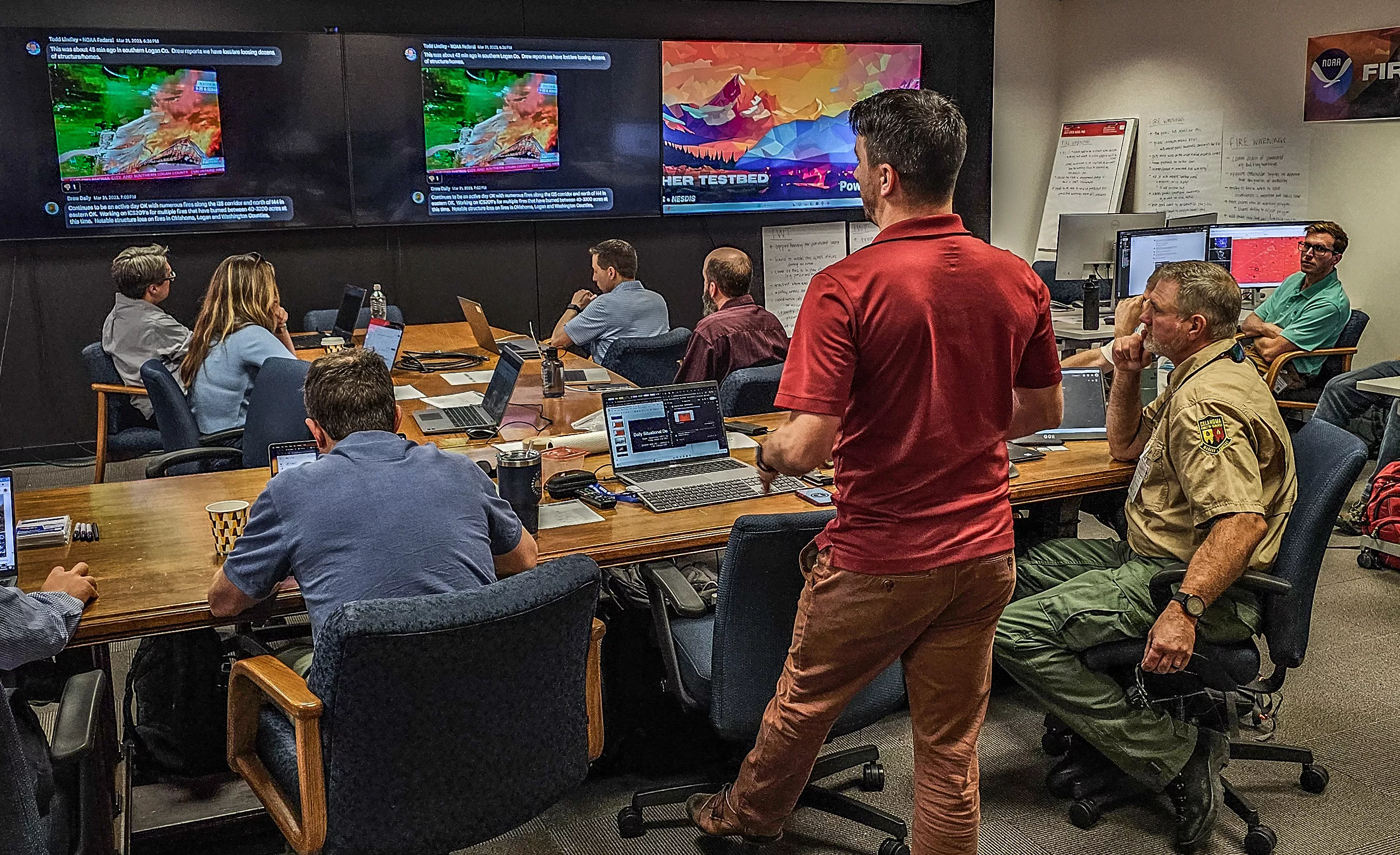 Kyle Thiem, a Test and Evaluation Meteorologist for the Fire Weather Testbed, provides a pre-brief in a conference room full of participants. The room has computer screens showing displaced real-time fire outbreak simulations, in which the participants practice detecting new wildfires and issuing Fire Warnings. 