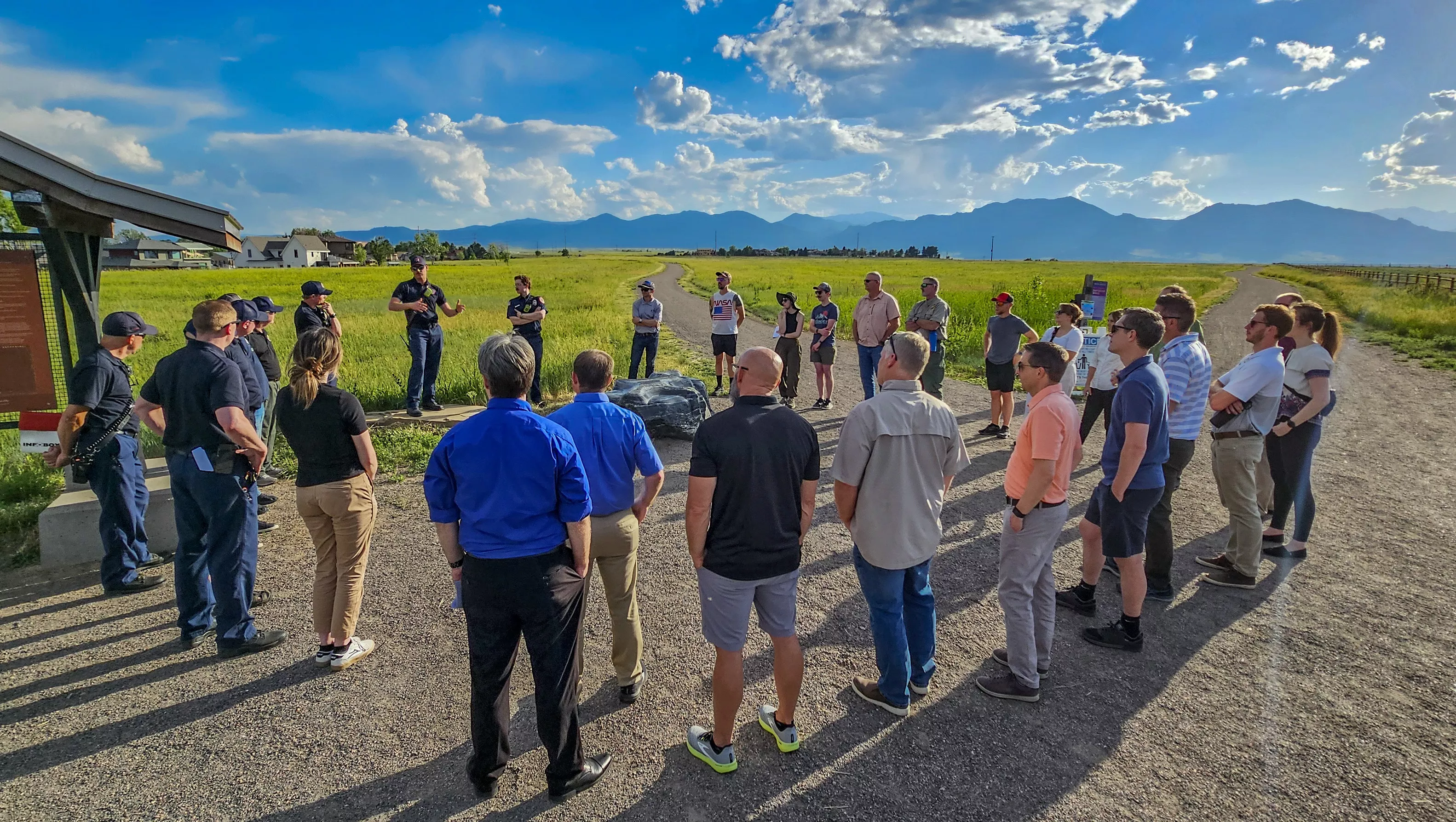 Firefighters in plain clothing from the Boulder, Louisville and Mountain View Fire Departments gather in a group on a sunny day outside in a dirt road in a field surrounded by mountains. They share their experiences battling the wind-driven Marshall Fire with researchers and testbed participants during a field trip.