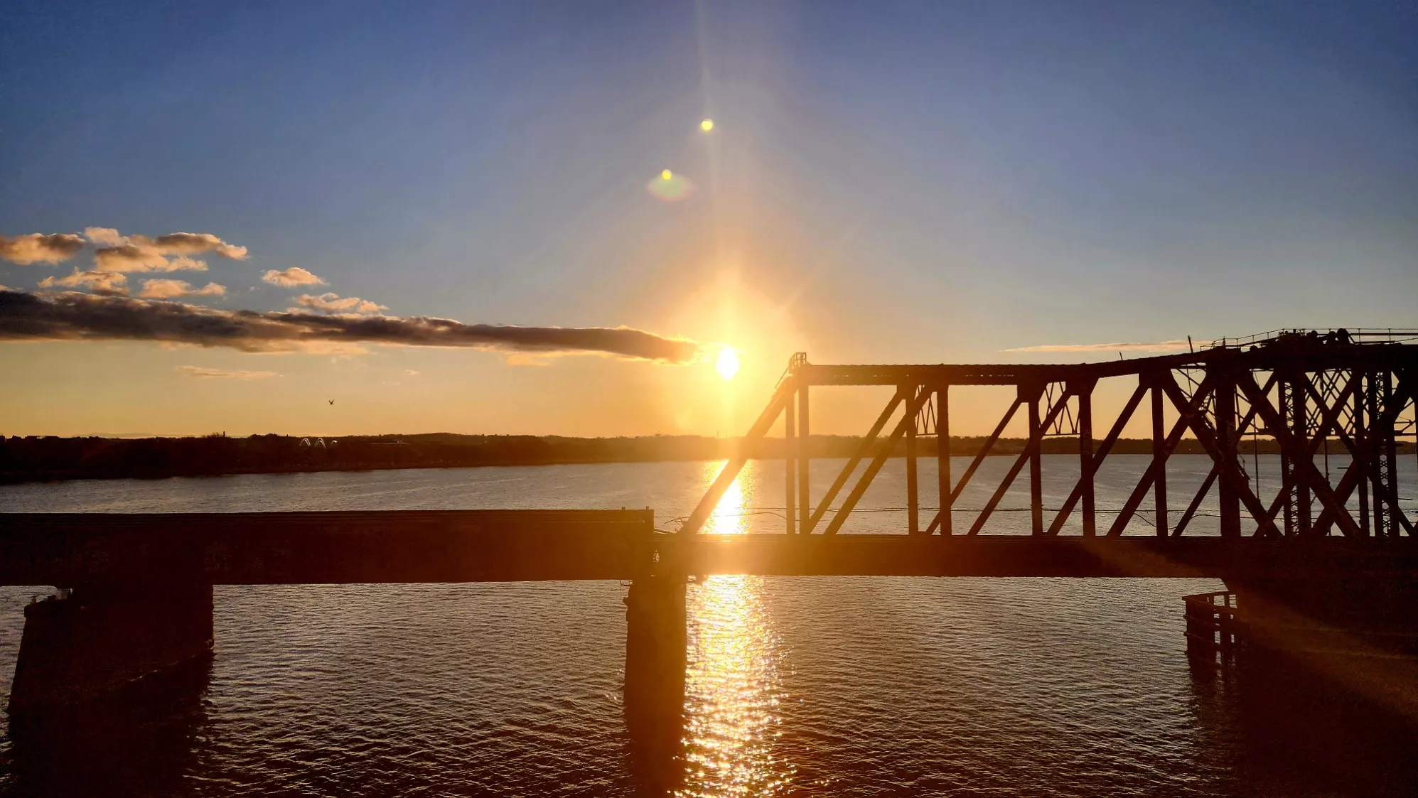 A sunrise over the Potomac River between Washington, D.C., and Virginia on the morning of the winter solstice. The golden sunlight reflects off the water, silhouetting a steel bridge against a clear sky with scattered clouds.