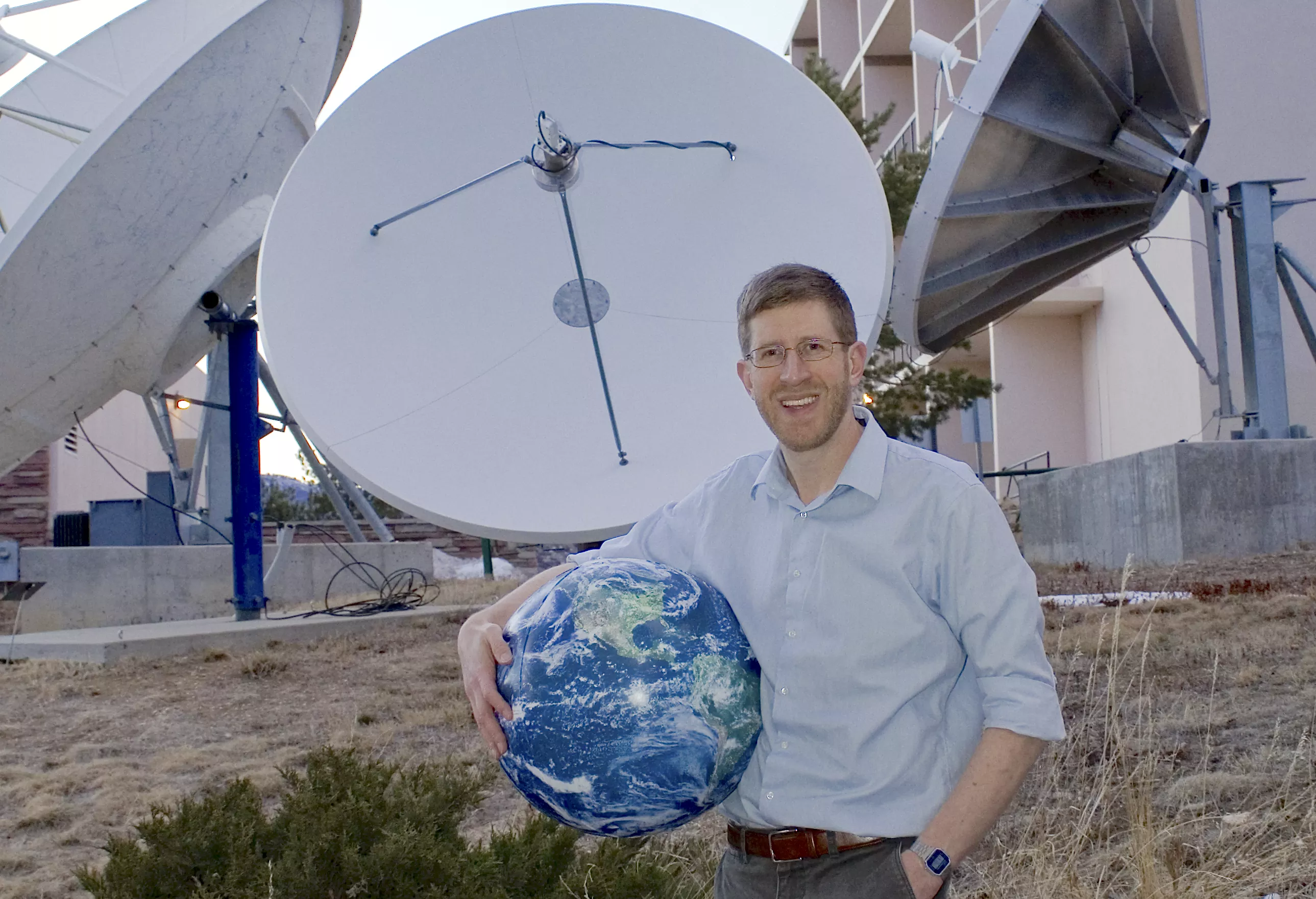 Chris Slocum standing in front of satellite dishes holding an inflatable Earth the size of a large beach ball.